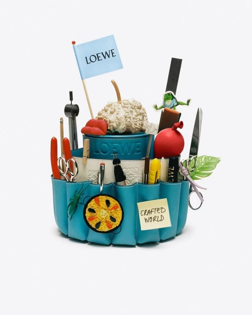 Craft supplies and various objects are arranged in a tiered blue container, with a Loewe flag and a 'Crafted World' sticky note.