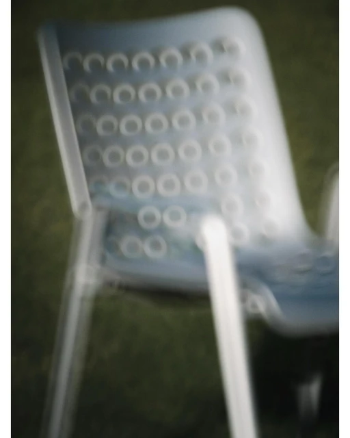 Close up of a blue plastic chair with a perforated back, casting shadows against an unfocused green background in blurred motion.