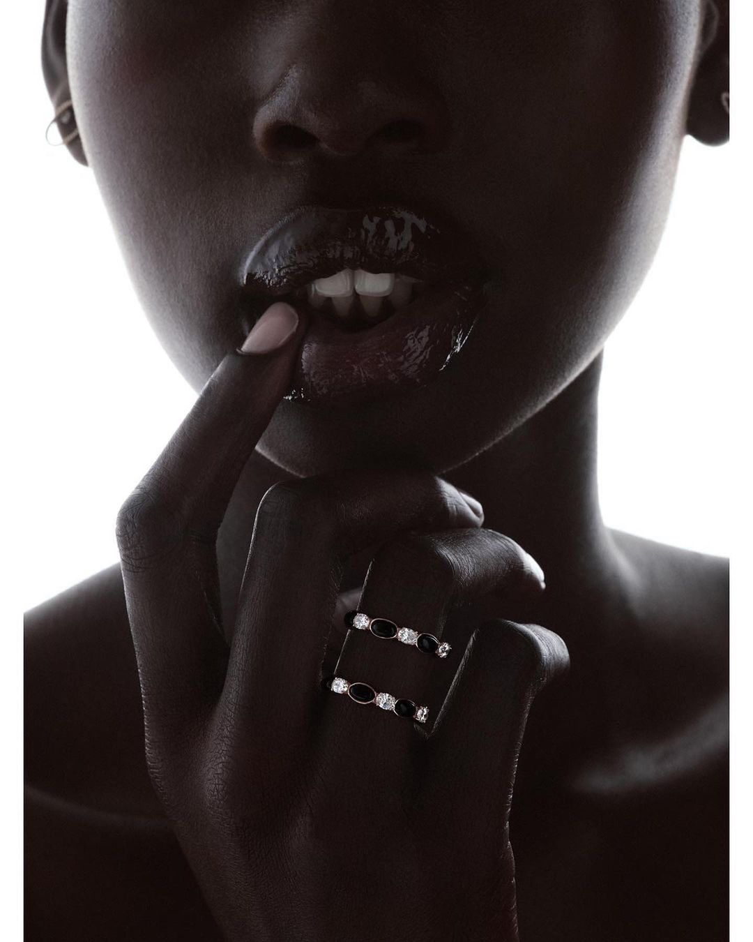 Close-up portrait of a black woman wearing two diamond and black gemstone rings, touching her glossy lips against a bright white background.