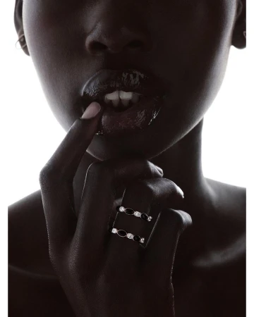 Close-up portrait of a black woman wearing two diamond and black gemstone rings, touching her glossy lips against a bright white background.