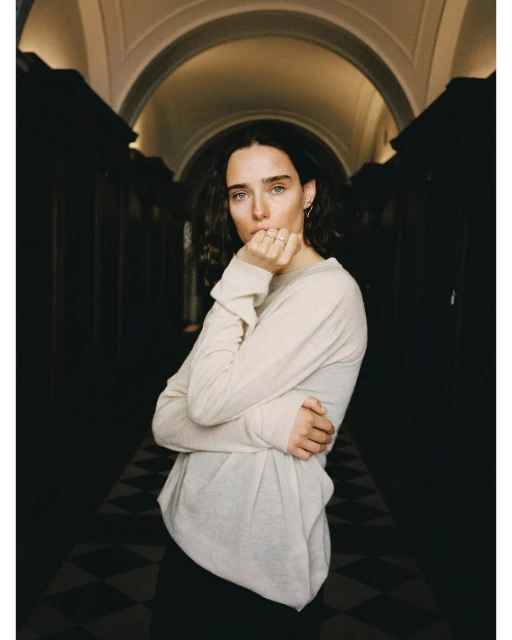 Indoor portrait of a young adult female with dark wavy hair, wearing a long-sleeved shirt, posing against a dark hallway backdrop with arched doorways.