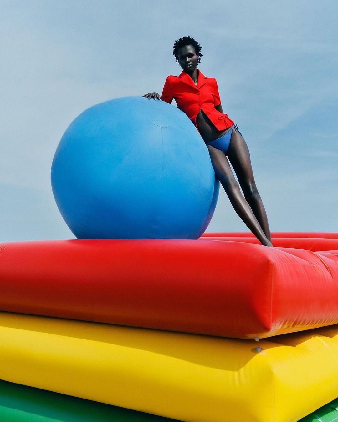 Adult woman in a red jacket poses with a blue ball on a stack of colorful inflatable platforms against a clear sky backdrop.