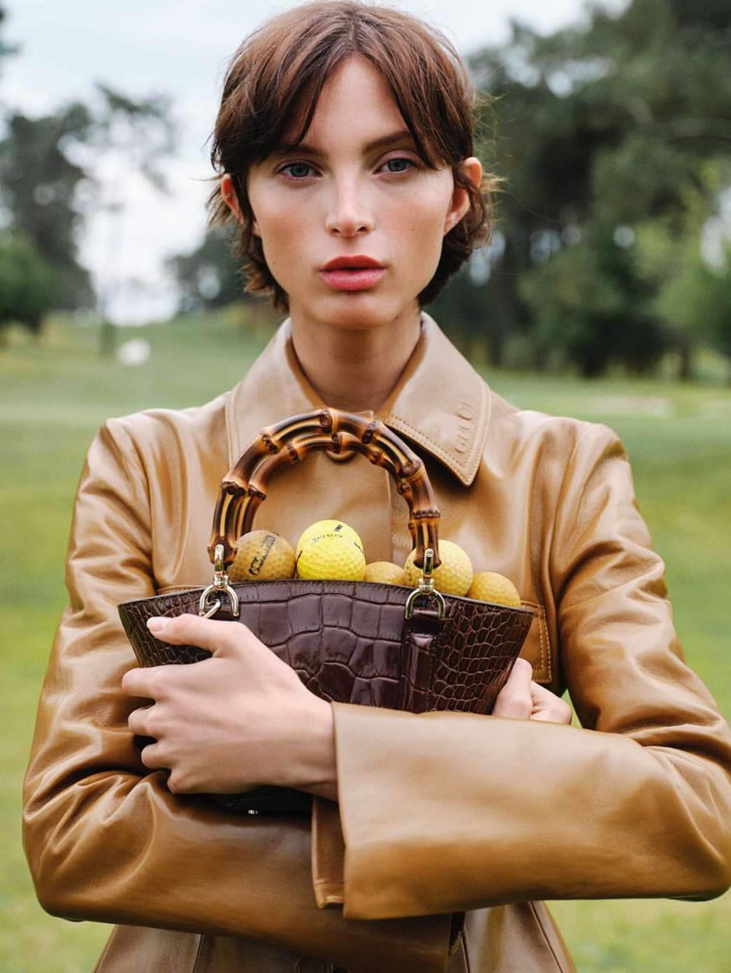 Brunette woman in a tan coat holds a brown, bamboo-handled purse filled with yellow golf balls against a green lawn and blurred trees.