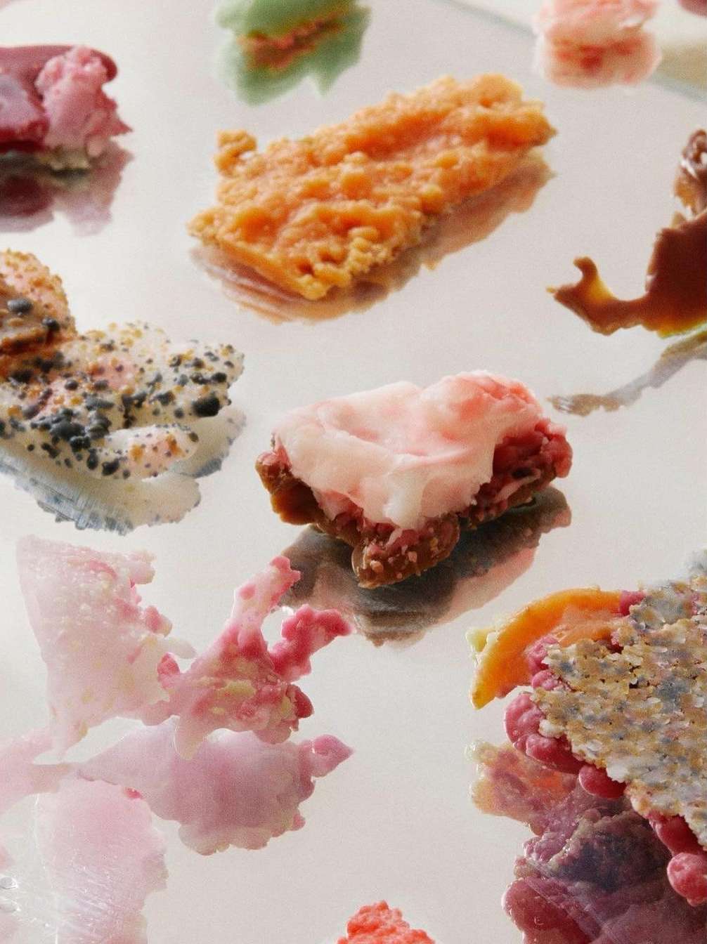 Overhead shot of colorful abstract shapes with rough textures, resembling coral or otherworldly delicacies, against a mirrored surface.