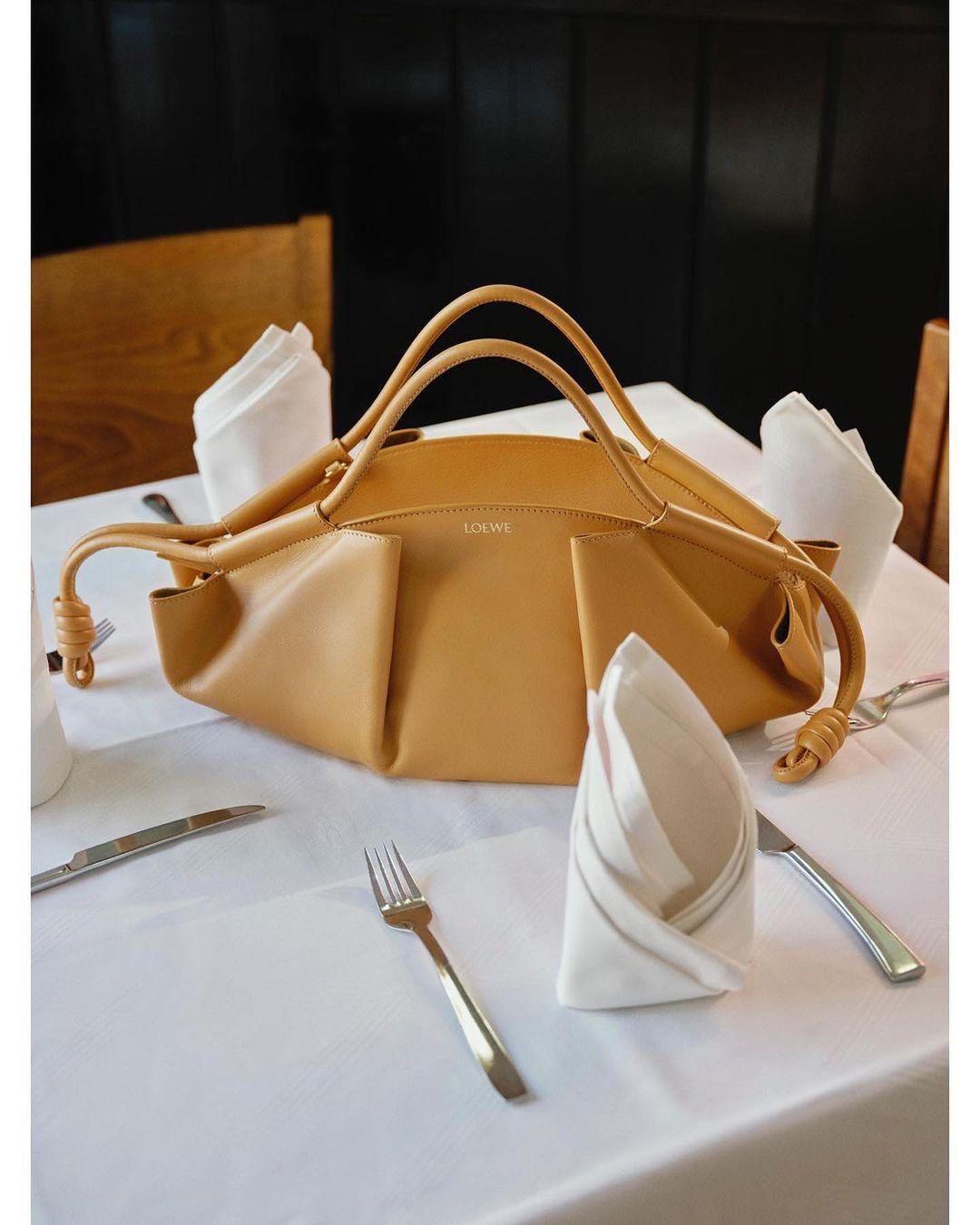 Tan Loewe handbag sits on a white tablecloth on a table with silverware and napkins, illuminated by natural light indoors.