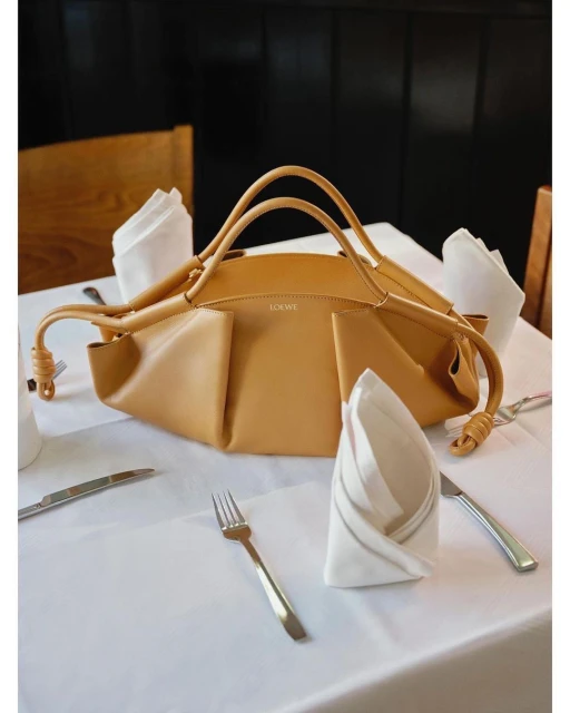 Tan Loewe handbag sits on a white tablecloth on a table with silverware and napkins, illuminated by natural light indoors.