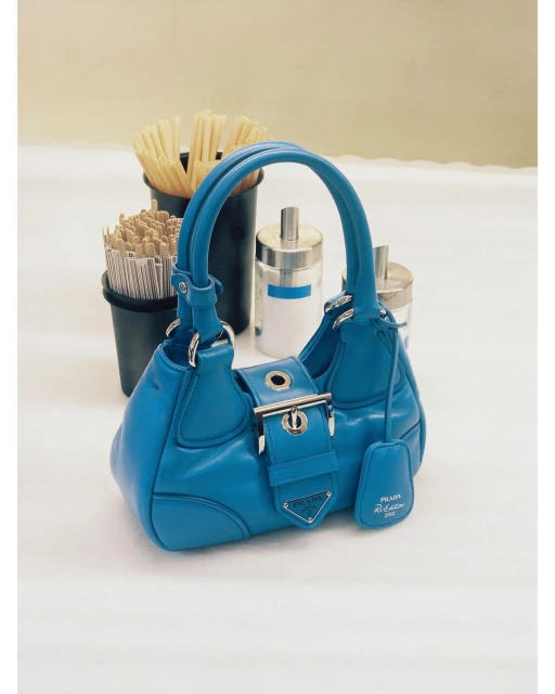 Turquoise Prada handbag with a silver buckle displayed on a white surface next to black containers holding small wooden sticks.