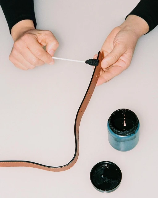 Close-up of hands applying black edge paint to the brown leather belt with an applicator next to an open jar of black paint.