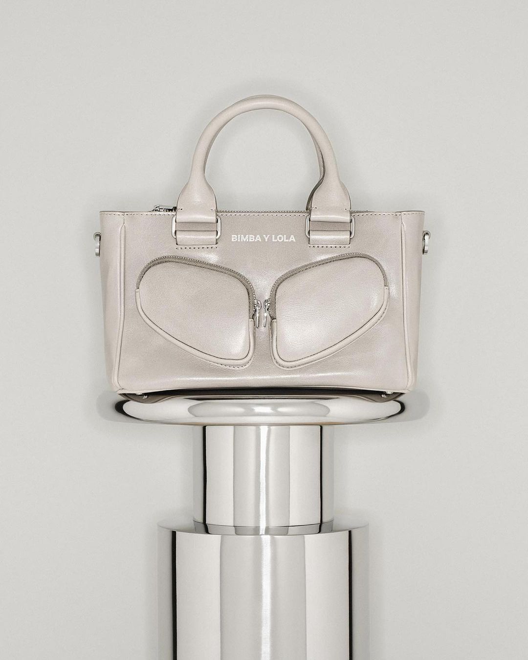 Beige leather tote bag by Bimba y Lola on a silver pedestal against a gradient gray background in a studio product shot.