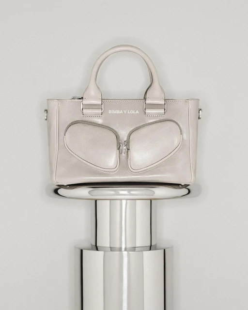 Beige leather tote bag by Bimba y Lola on a silver pedestal against a gradient gray background in a studio product shot.
