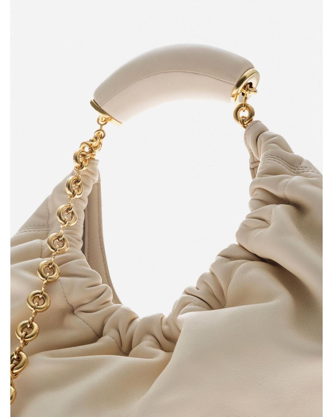 Close-up of a beige leather hobo bag with a gold chain strap and a curved handle, set against a bright white background, fashion accessory.