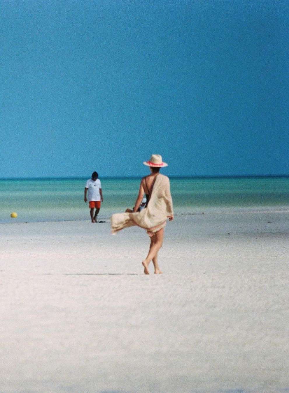 Woman in swimwear and cover-up walking on a beach with a man in the background under a clear blue sky and turquoise water.