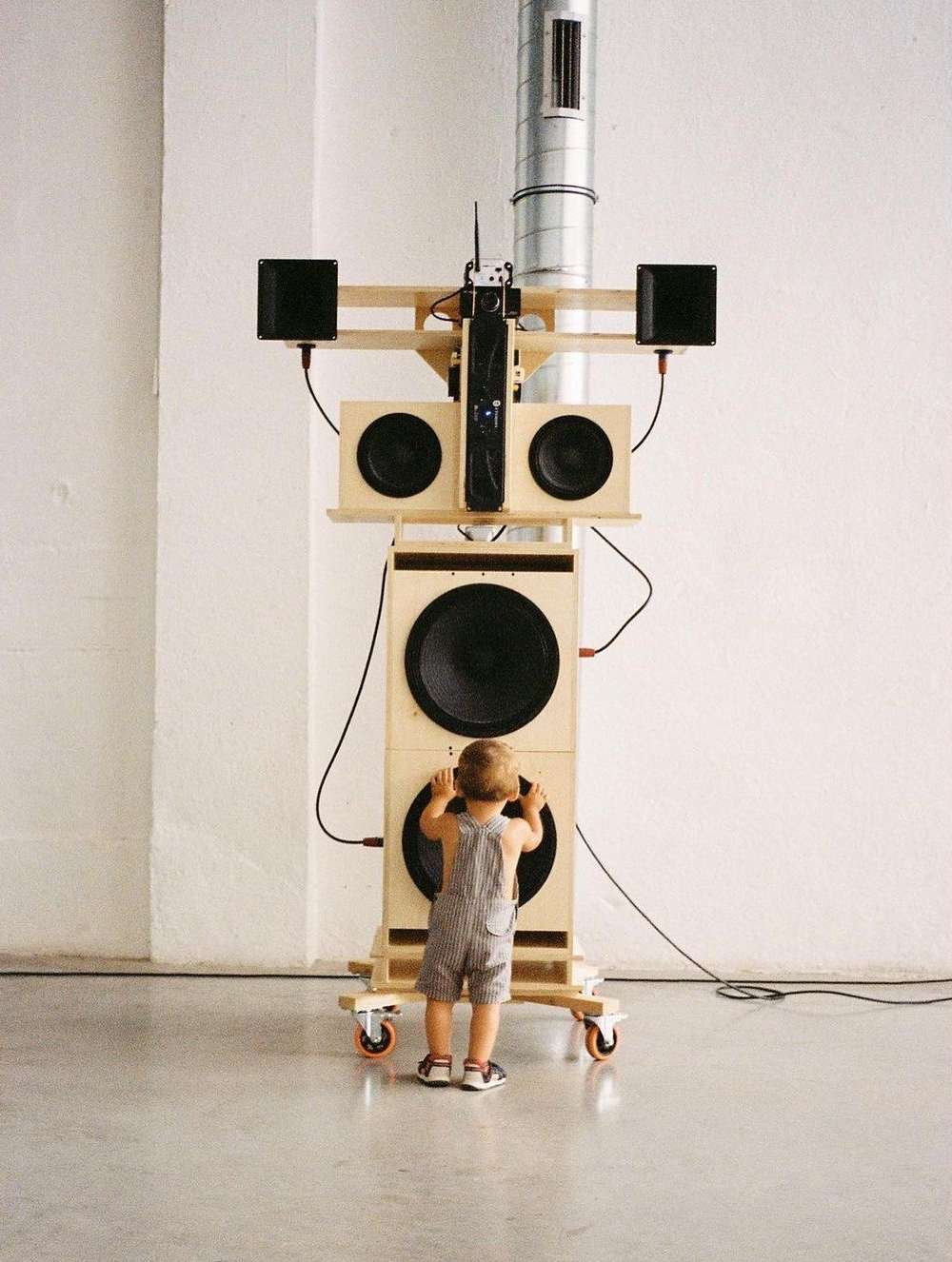 Young child stands in front of tall custom speaker setup with speakers and electronics, with grey concrete floor and white wall.