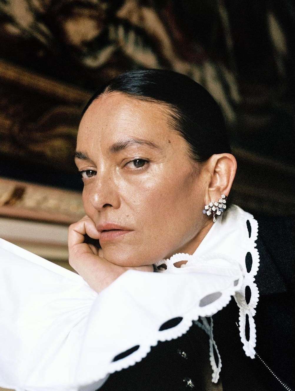 Close-up portrait of a woman with her hand on her cheek, wearing a black top with a white collar, against a blurred background.