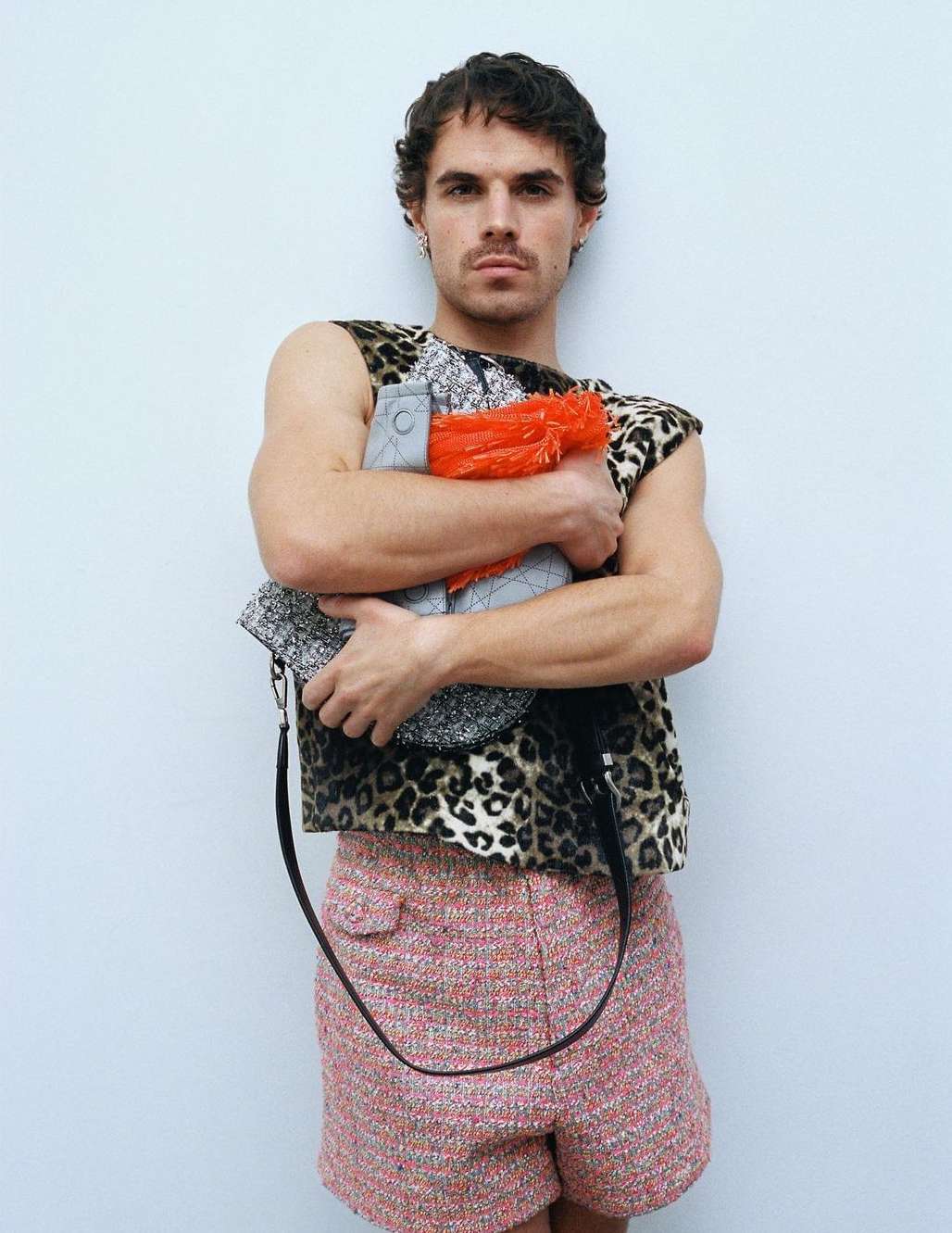 Adult male with short curly hair poses in animal print top, red and grey bag, and patterned shorts against a light grey wall.