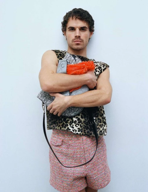 Adult male with short curly hair poses in animal print top, red and grey bag, and patterned shorts against a light grey wall.