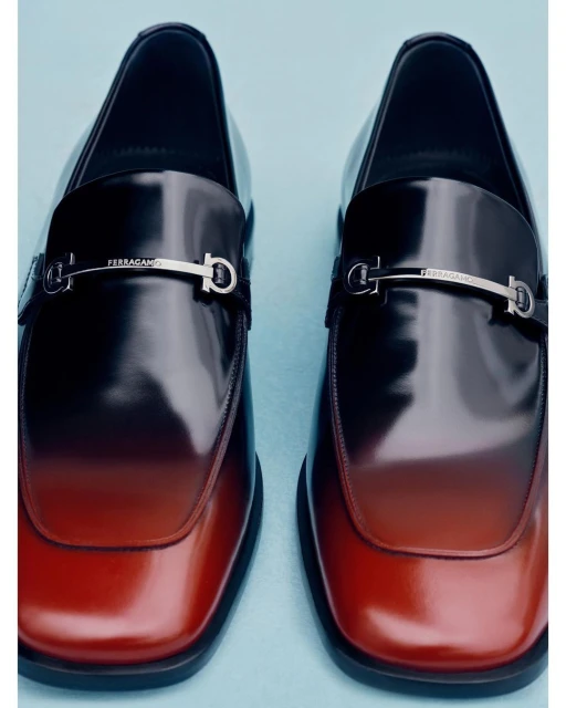 A pair of sleek, two-tone leather loafers, featuring a smooth gradient finish from dark brown to reddish-brown, and polished silver hardware.
