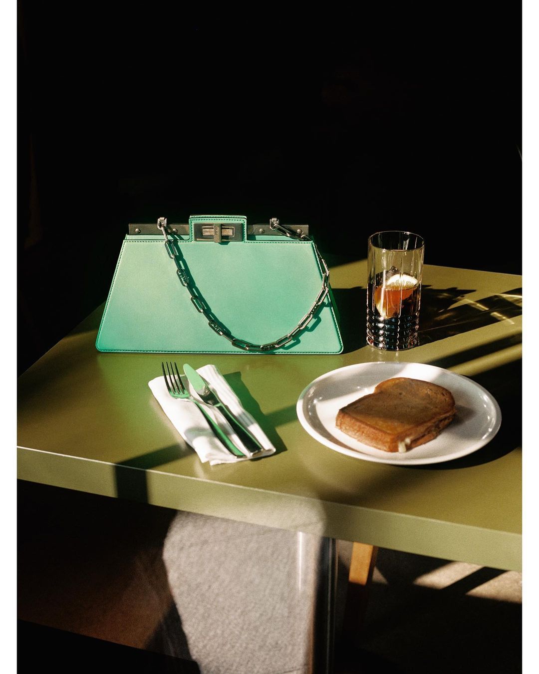 Stylish mint green satchel handbag displayed on a table next to a water glass, sandwich, and silverware, in a dimly lit setting.