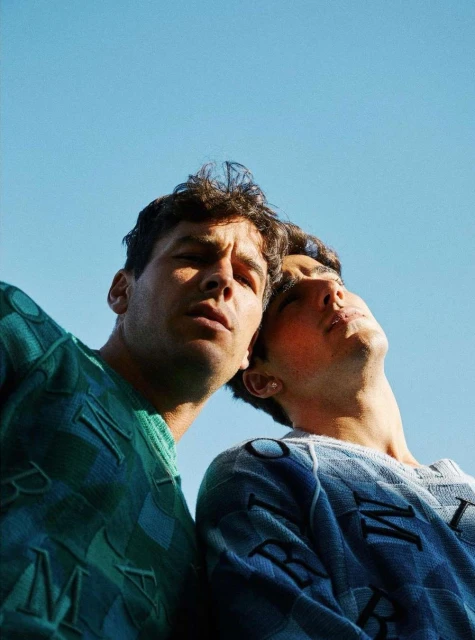 Two young men wearing patterned sweaters pose in a low-angle shot with a clear blue sky as the backdrop, creating a striking fashion image.