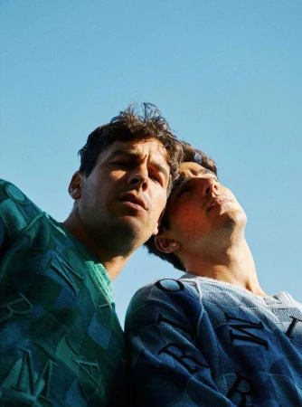 Two young men wearing patterned sweaters pose in a low-angle shot with a clear blue sky as the backdrop, creating a striking fashion image.