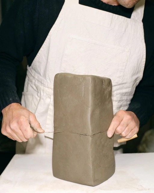 Close-up of a person cutting a block of clay with a wire tool on a white surface, wearing a white apron, showing hands and clay.