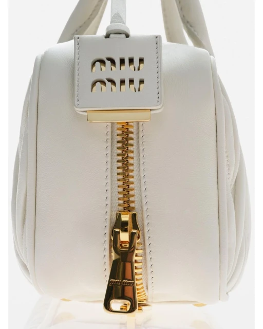 Close-up studio shot of a white leather Miu Miu handbag with a gold zipper and logo tag. Fashion product photography, isolated on white.