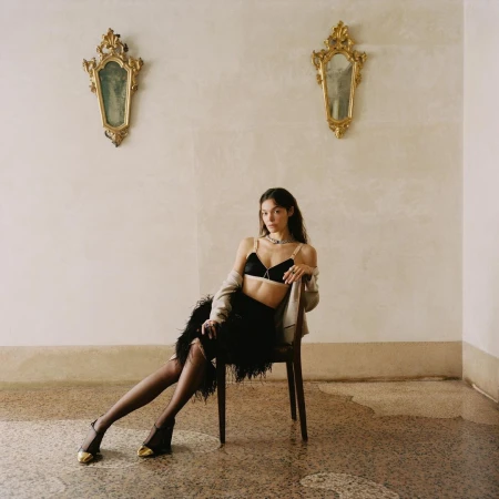 Woman in black bra and feather skirt seated on a dark wood chair in a neutral room with gold mirrors