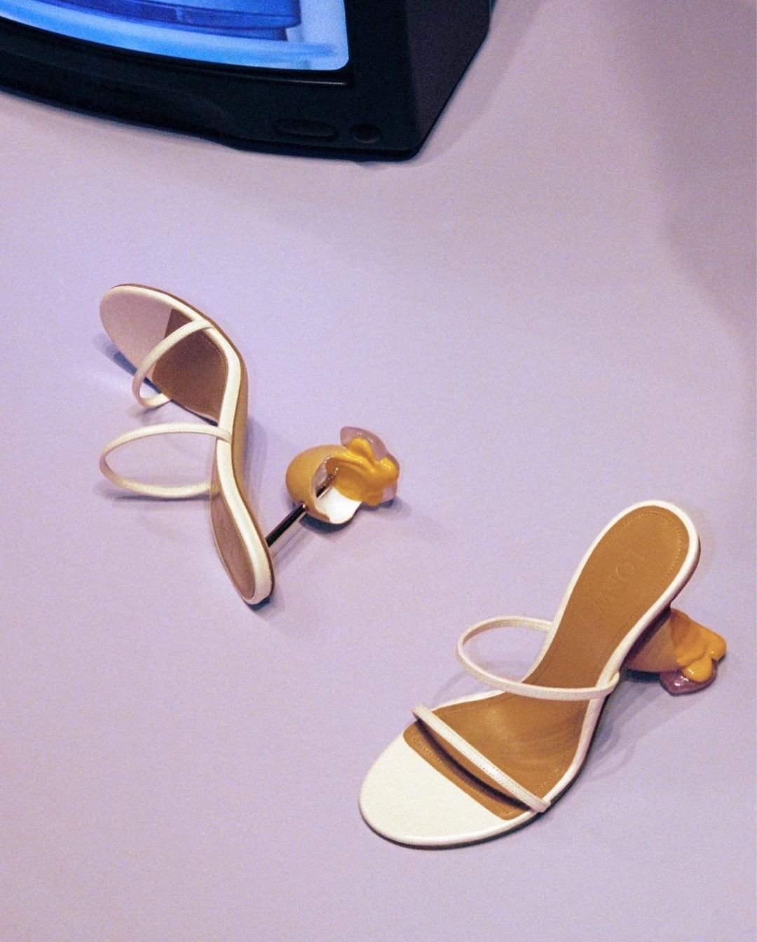 Two stylish white strappy heeled sandals are showcased on a lilac surface next to a vintage television set.