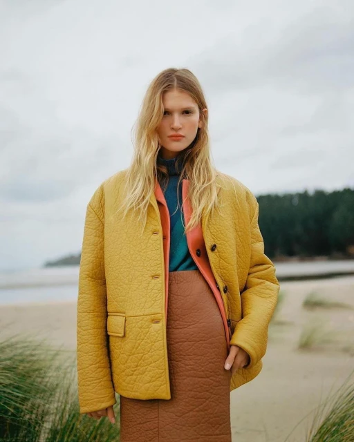 An adult woman stands on a beach modelling a yellow quilted jacket and brown skirt. The background shows sand, dunes, and a grey sky.