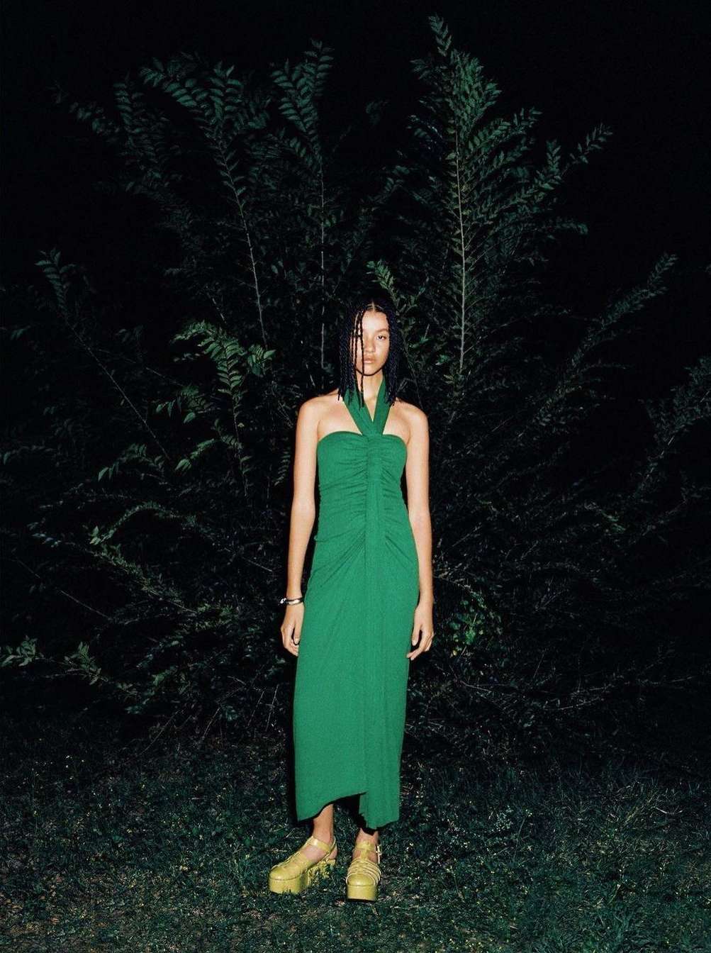Woman in a green halter dress and platform sandals stands in front of dark green foliage in a dark outdoor setting.