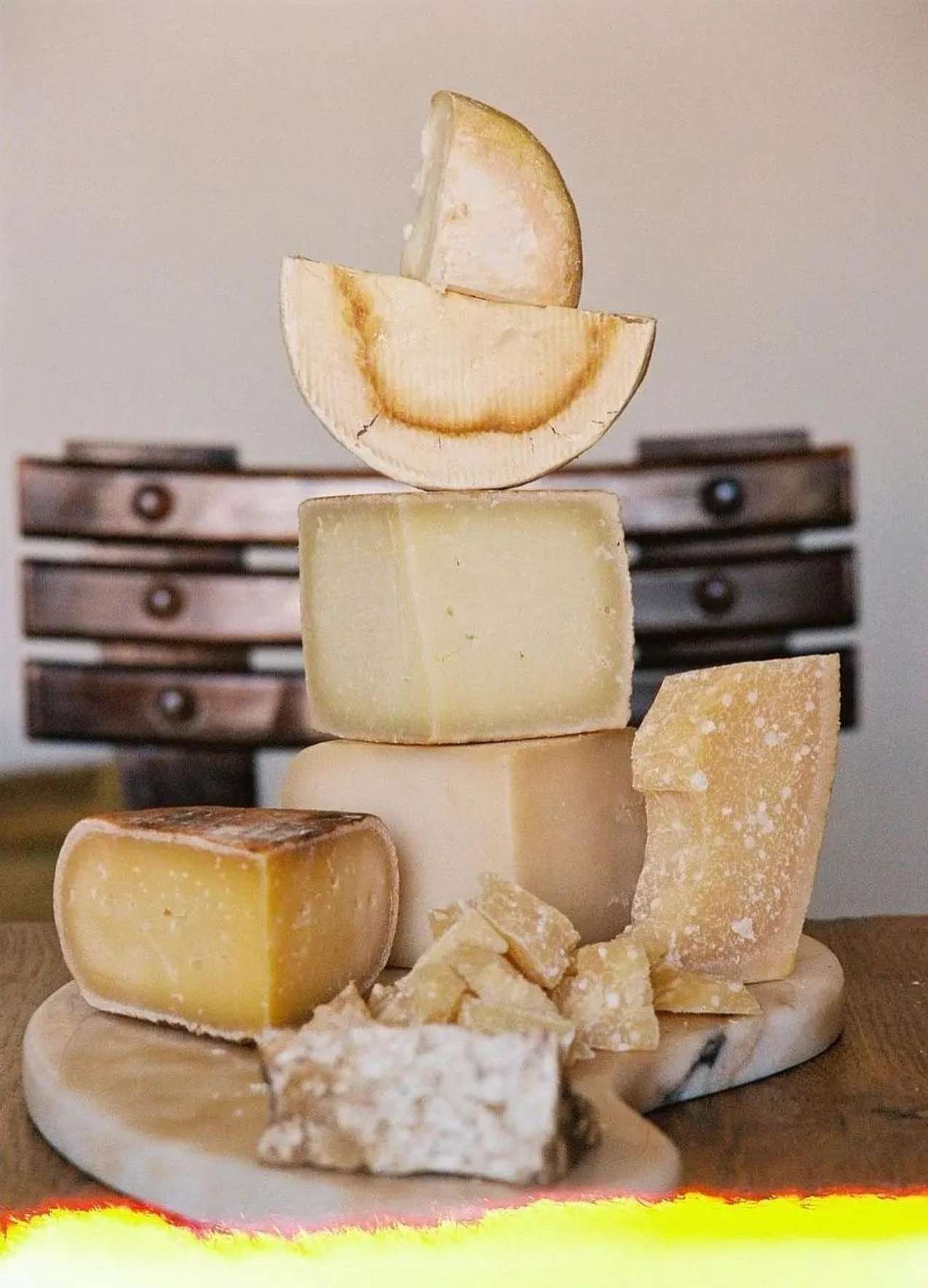 Stacked cheese blocks and wedges of various textures and colours on a round marble board with a wooden rack visible in the background.