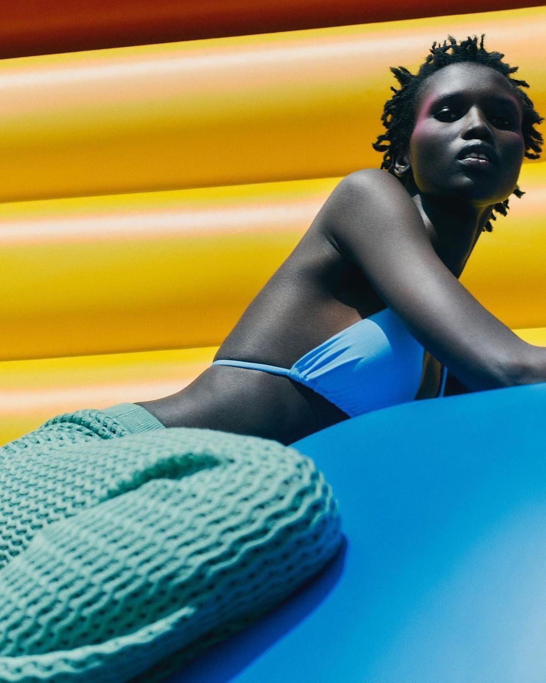 Adult female model with dark skin poses in a light blue bikini top and knitted green pants against a bright yellow and blue background.
