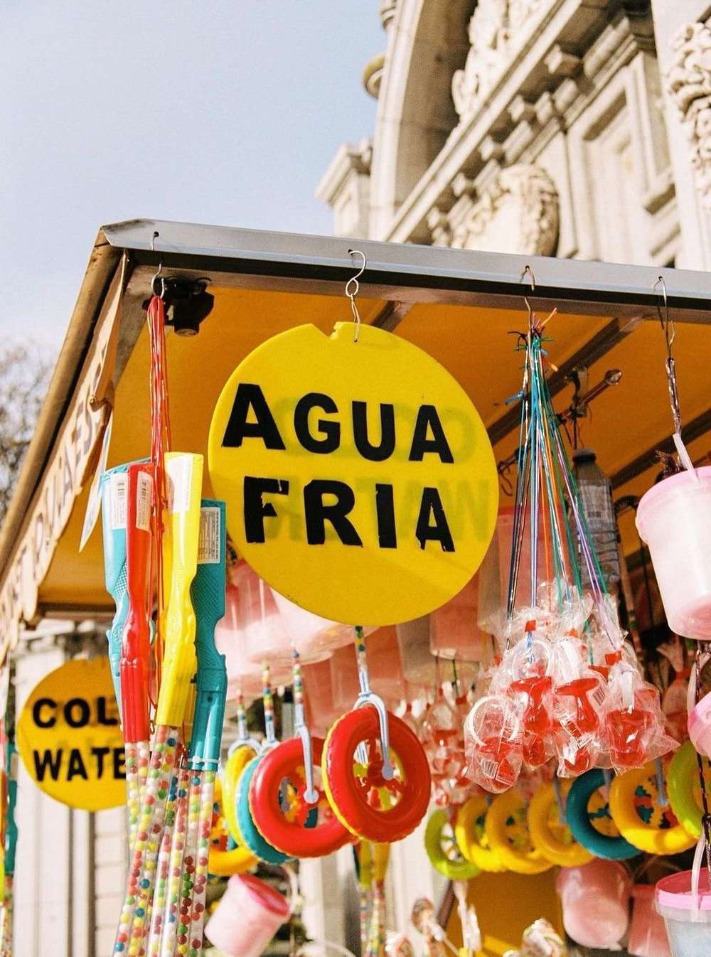 Yellow market stand displays signs for 'Agua Fria' (Cold Water) along with colorful toys and treats, set against a building facade.