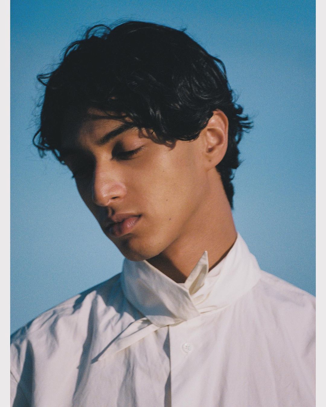 Portrait of a man with dark wavy hair wearing a white collared shirt against a clear blue sky background.