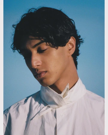 Portrait of a man with dark wavy hair wearing a white collared shirt against a clear blue sky background.