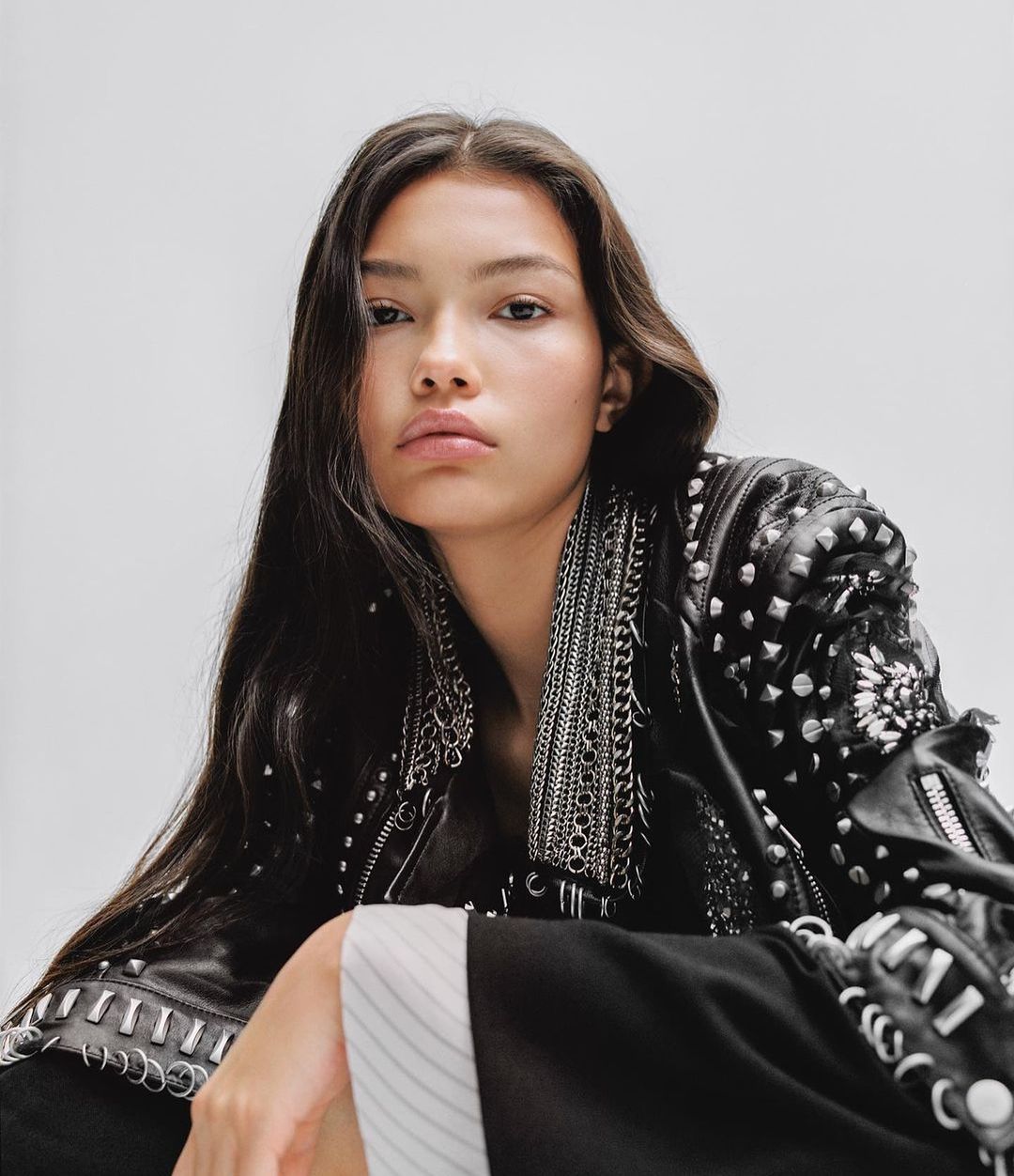 A young woman with long brunette hair poses in a studded black leather jacket against a clean white background in a high-angle studio shot.
