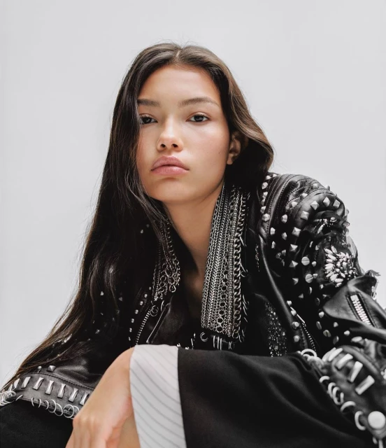 A young woman with long brunette hair poses in a studded black leather jacket against a clean white background in a high-angle studio shot.