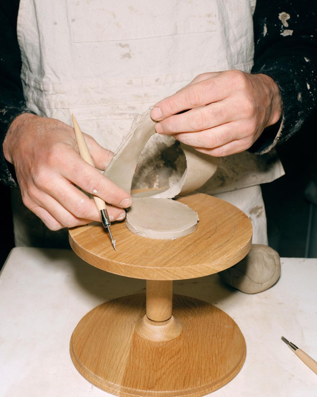 Adult hands using a sculpting tool to shape a piece of clay on a wooden pottery wheel in a workshop setting. Clay shaping process.