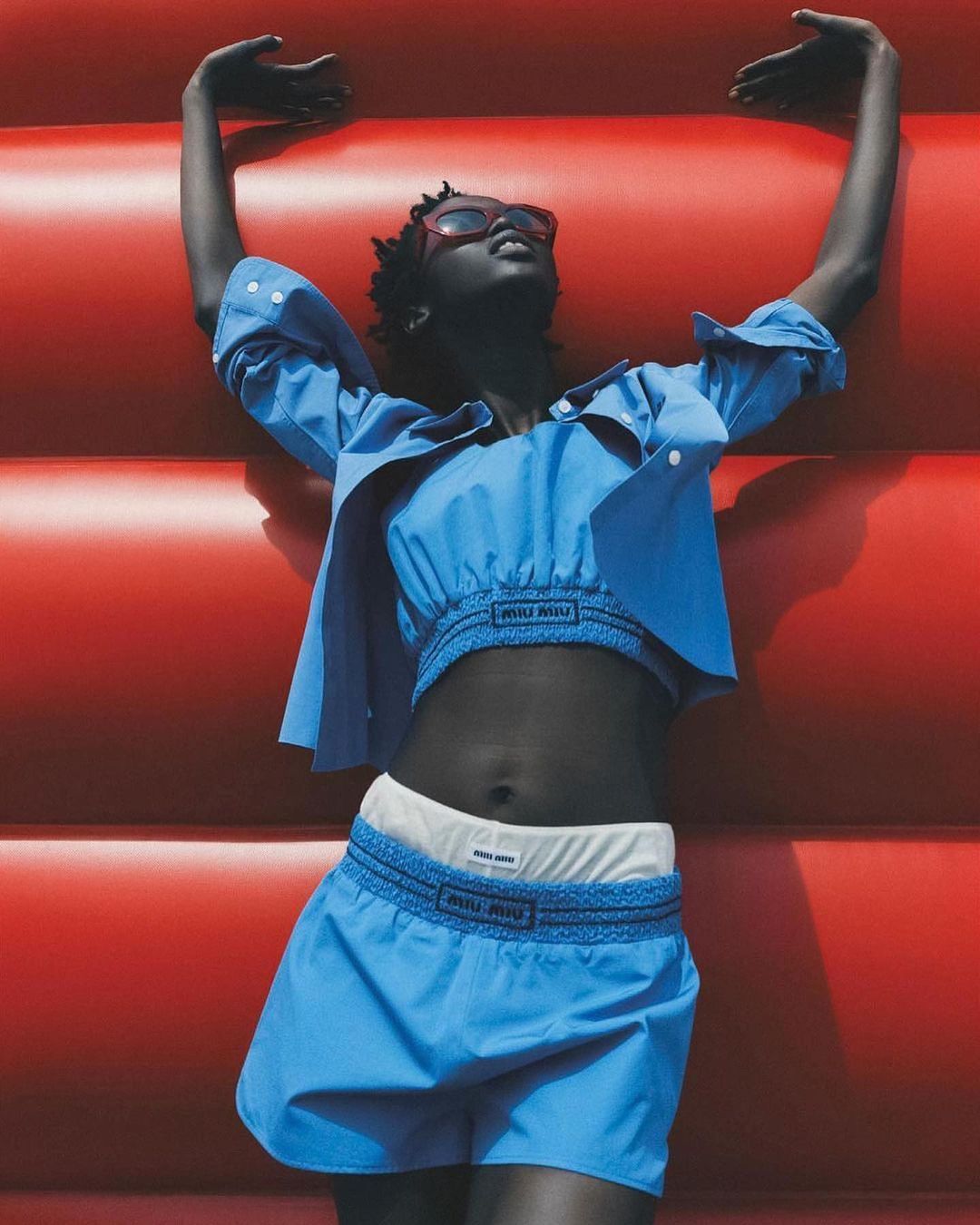 A Black female model with a short Afro hairstyle poses against a red wall, wearing a blue Miu Miu crop top, shirt, and shorts with white underwear.