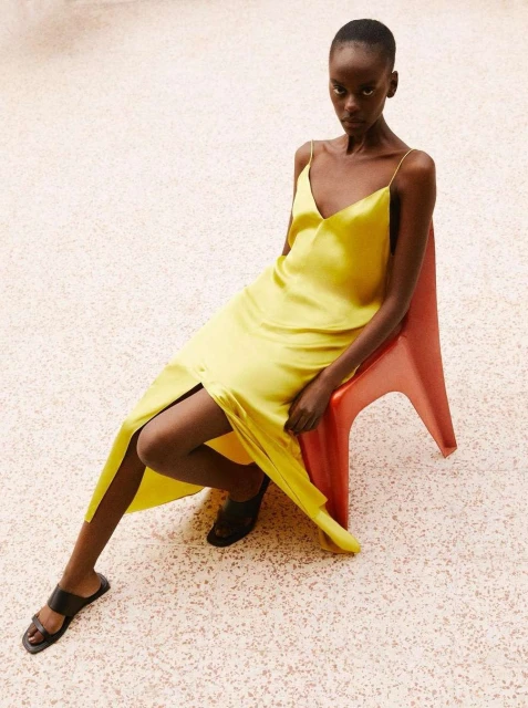 A Black woman poses while seated on a red plastic chair in a studio. She is wearing a yellow silk dress and black sandals.