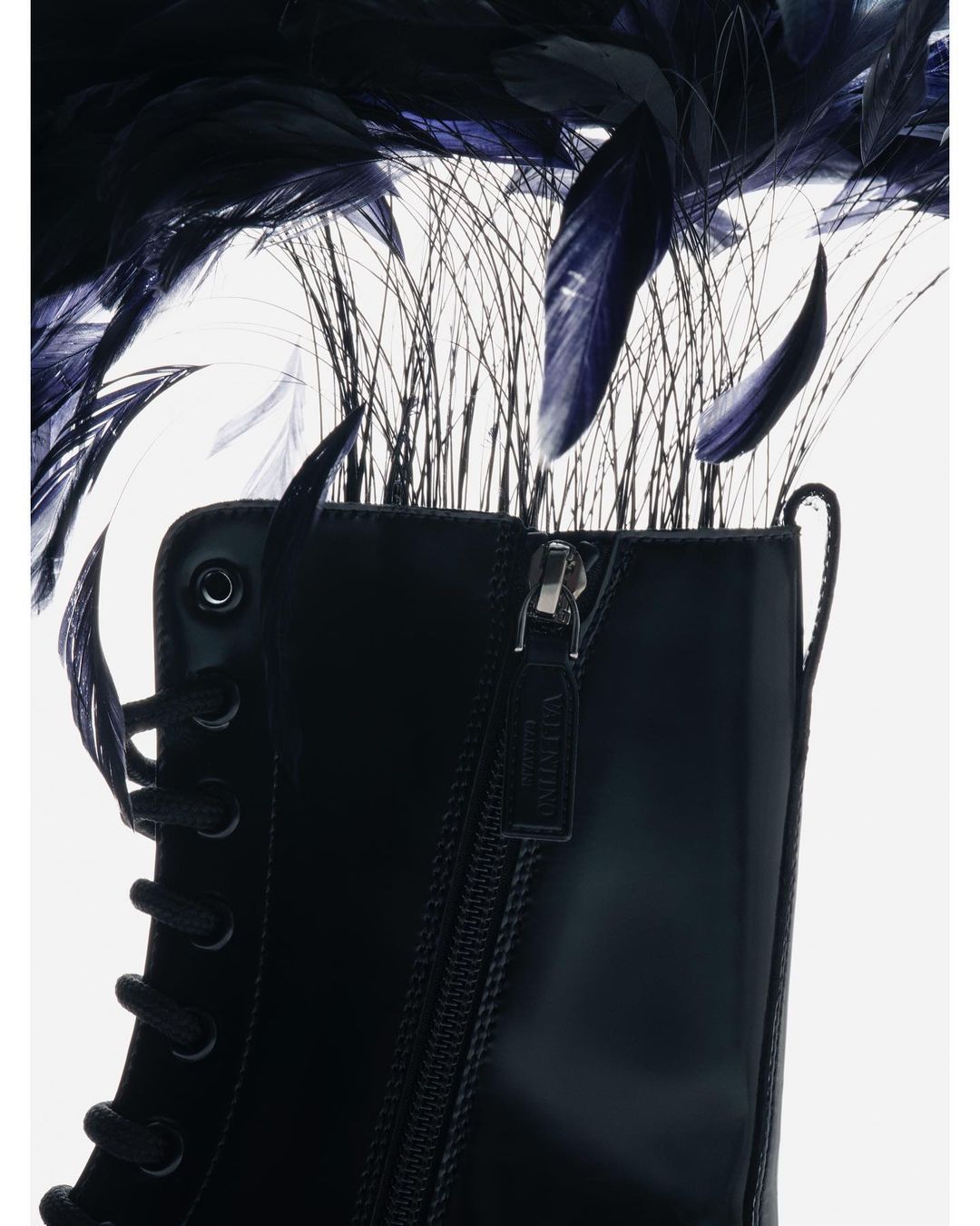 Black lace-up boot close-up against a white backdrop adorned with black feathers and thin grass-like decorations above the boot.