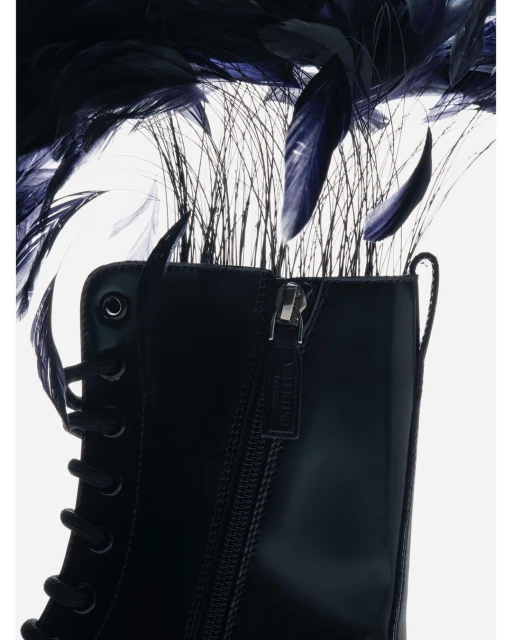 Black lace-up boot close-up against a white backdrop adorned with black feathers and thin grass-like decorations above the boot.