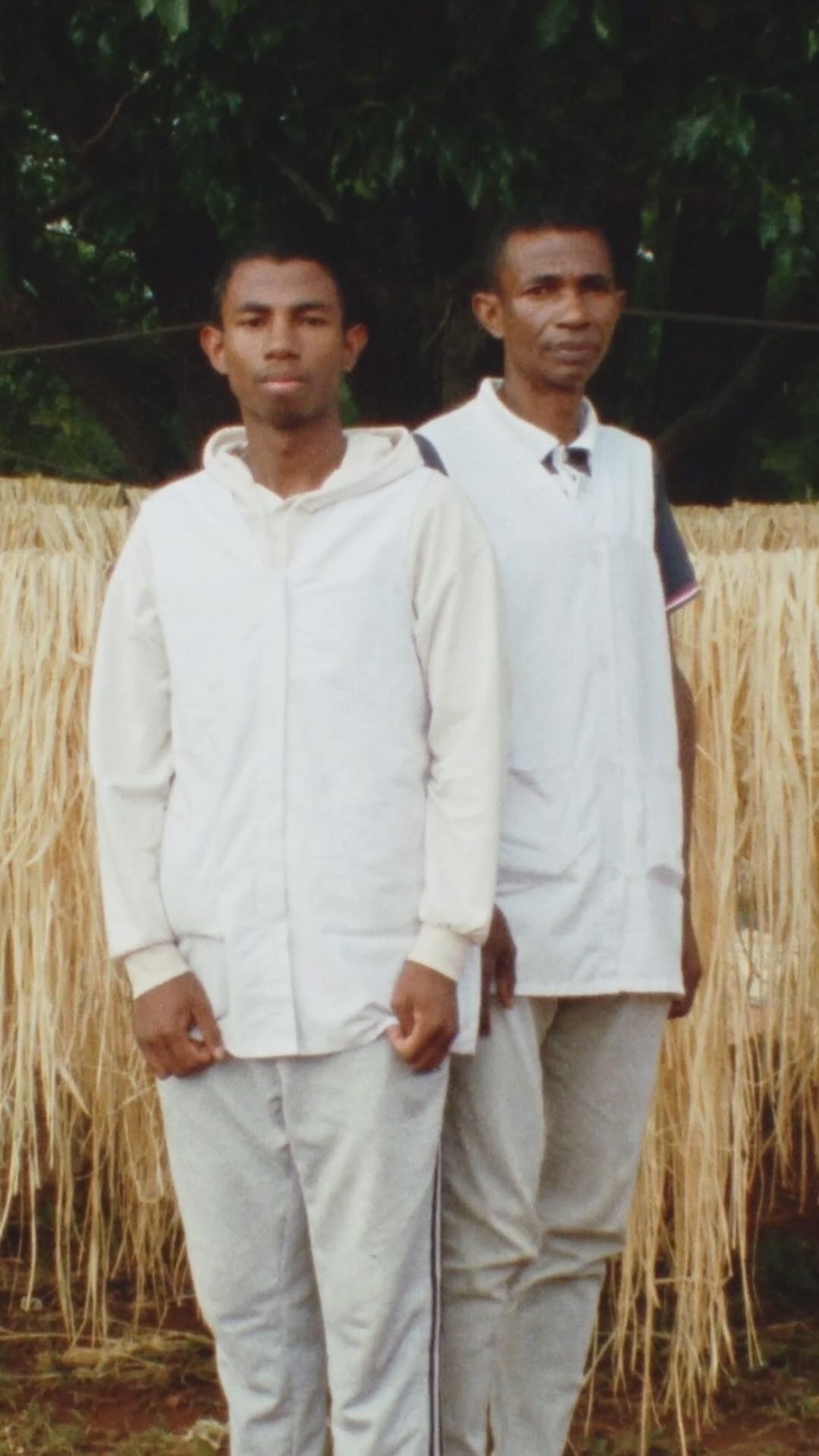 Two adult men stand outdoors in casual clothing against a backdrop of reeds and greenery. One wears a white vest, the other a hooded top.