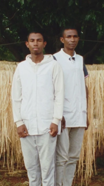 Two adult men stand outdoors in casual clothing against a backdrop of reeds and greenery. One wears a white vest, the other a hooded top.