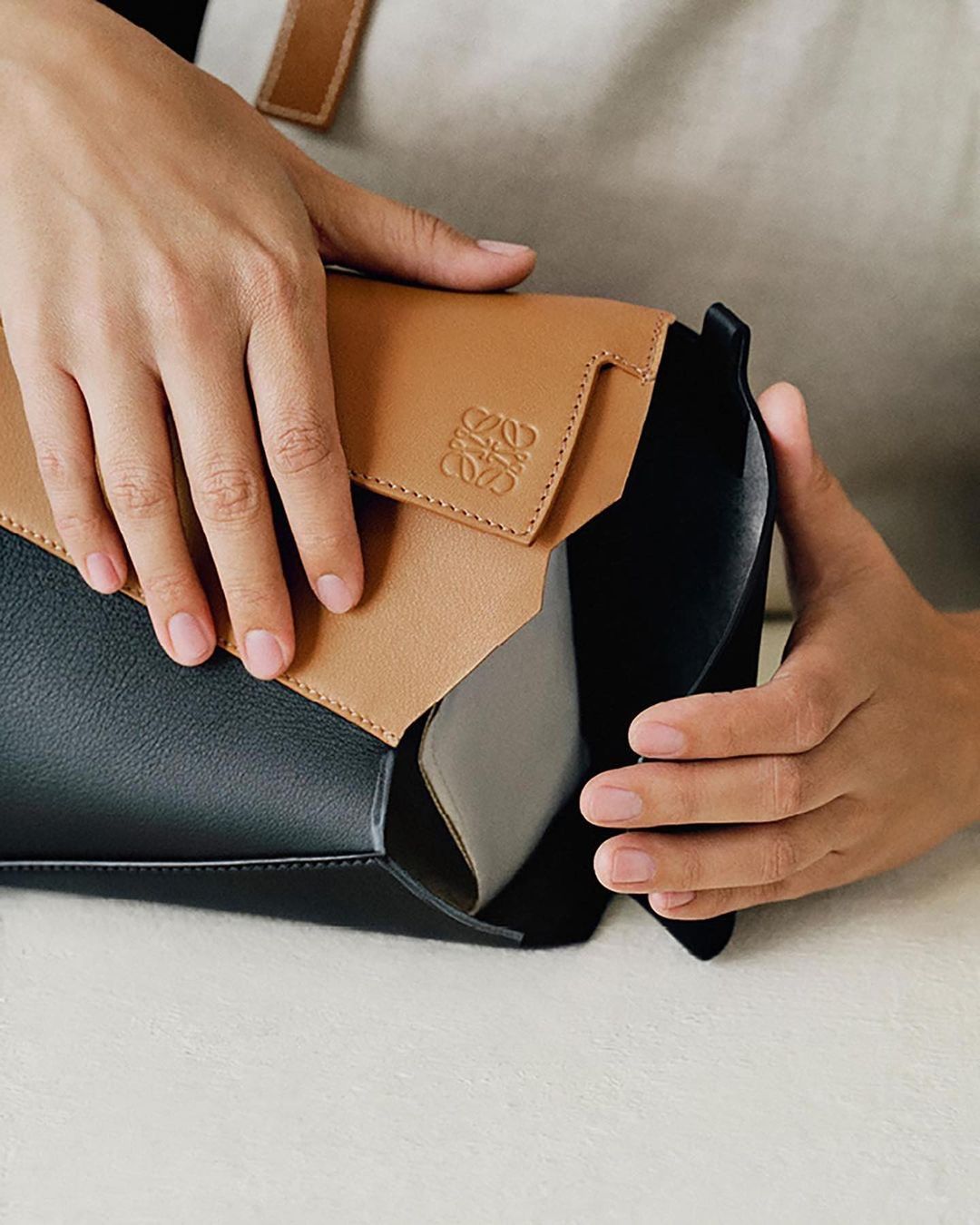A person's hands opening a black and tan leather handbag. The bag has a tan flap with a repeating symbol on it. The interior is grey.