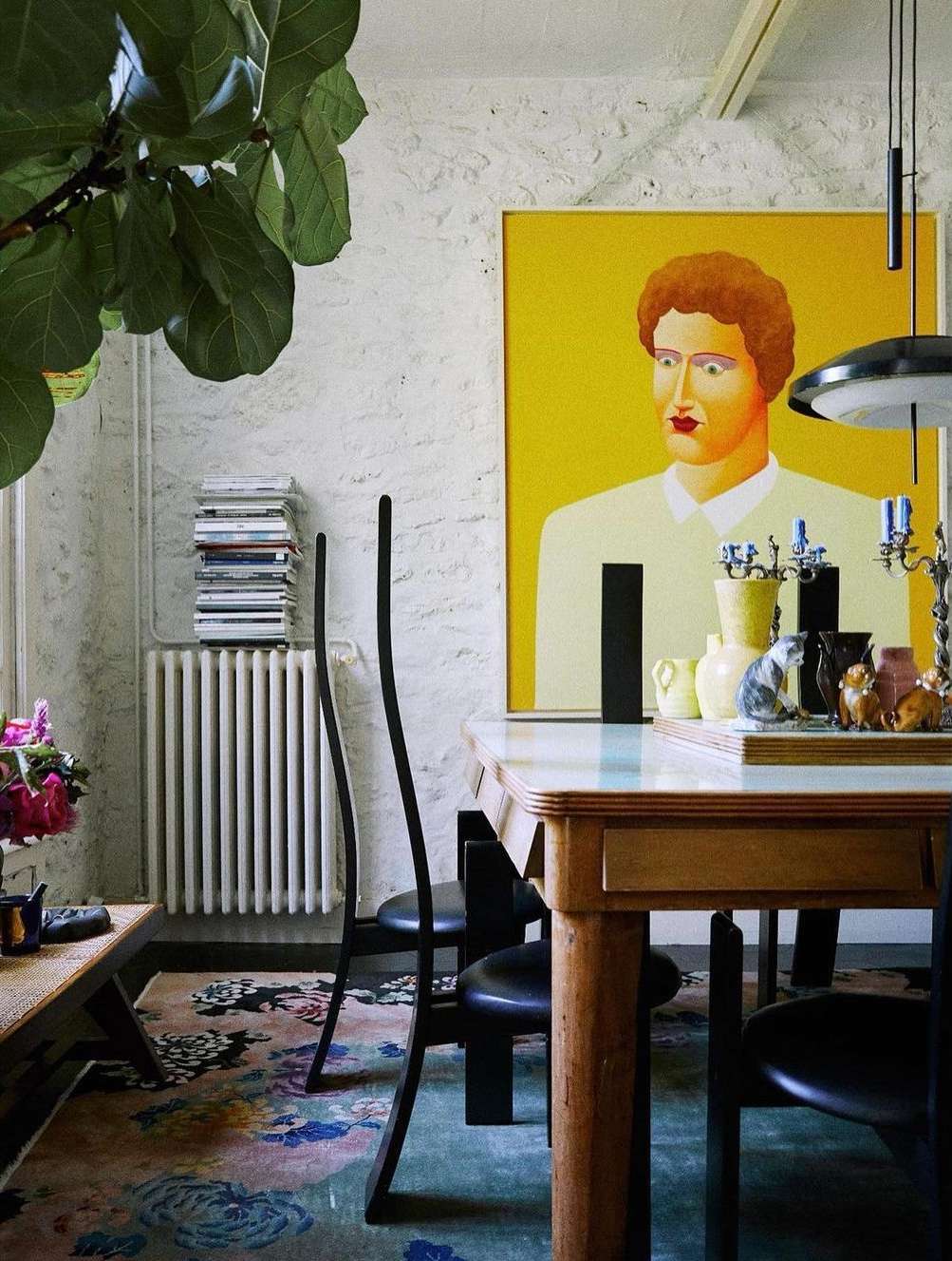 Bright yellow wall art depicting a person hangs over a dining table with black chairs in a modern interior, alongside a radiator and greenery.
