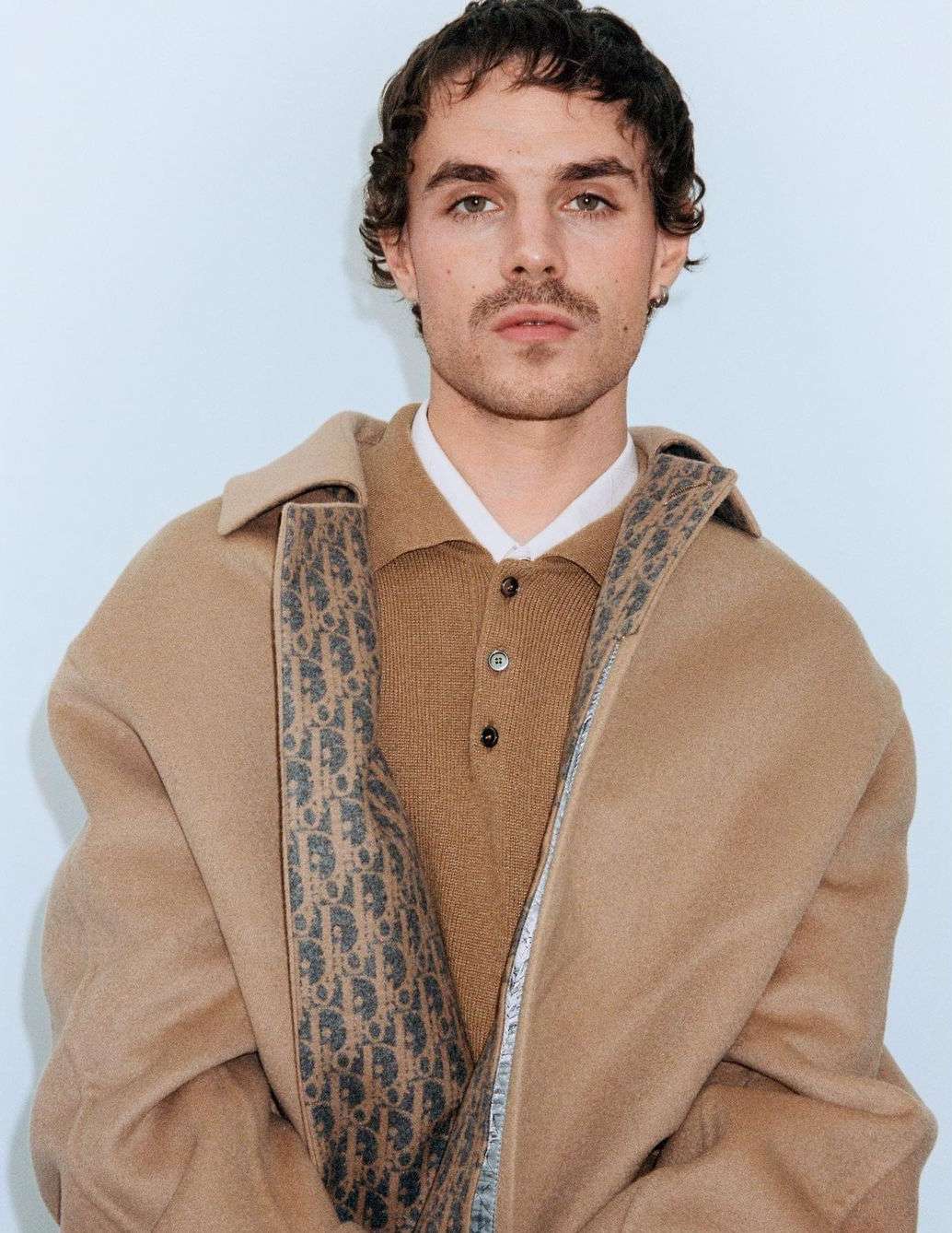 A portrait shot of a young adult male with brown hair wearing a brown collared shirt and a beige coat with a logo pattern on the lapels.