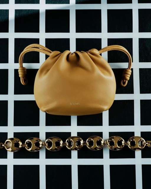 Tan Loewe pouch bag and gold chain necklace on a black and white grid background, showcasing fashion accessories in a studio setting.