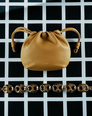 Tan Loewe pouch bag and gold chain necklace on a black and white grid background, showcasing fashion accessories in a studio setting.