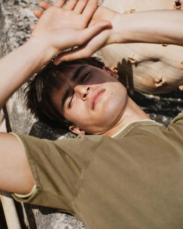 Young man lying down outdoors with hands raised above his head, wearing an olive green t-shirt under bright sunlight.
